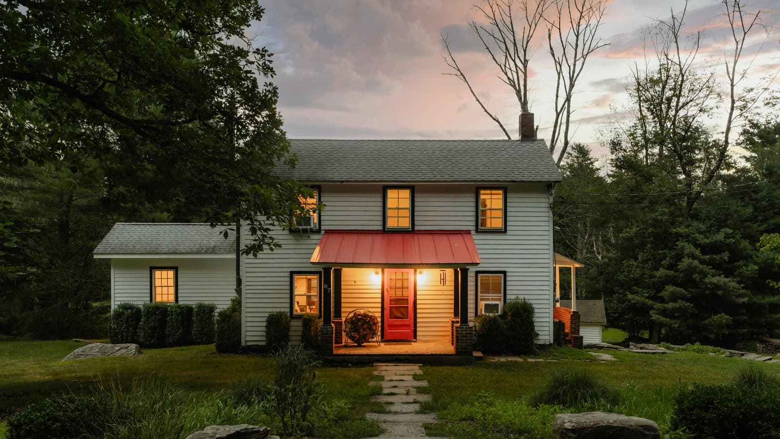 A white two-storey farmhouse at dusk with the porch light on and warm light in the windows.